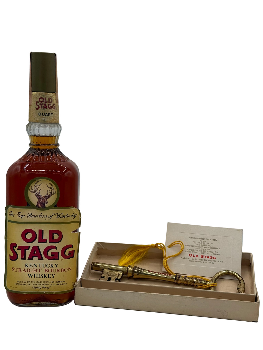 Photograph of a vintage Old Stagg Kentucky Straight Bourbon Whiskey bottle from Frankfort, KY, featuring a stag emblem and an Eighty Proof label, displayed with a brass-toned commemorative key with yellow tassel celebrating the 2,000,000th barrel from the Albert B Blanton Distillery