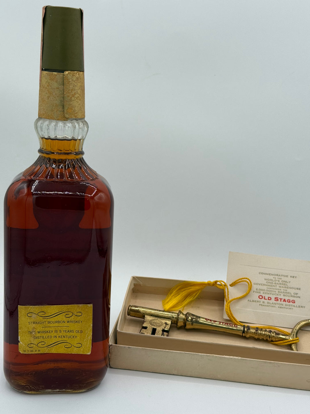 A vintage Old Stagg Straight Bourbon Whiskey bottle containing amber liquid with a yellow label is displayed alongside a gold commemorative key with a yellow tassel and an informational card detailing the World's Only One-Barrel Government Warehouse and the 2,000,000th barrel from the Albert B Blanton Distillery in Frankfort Kentucky