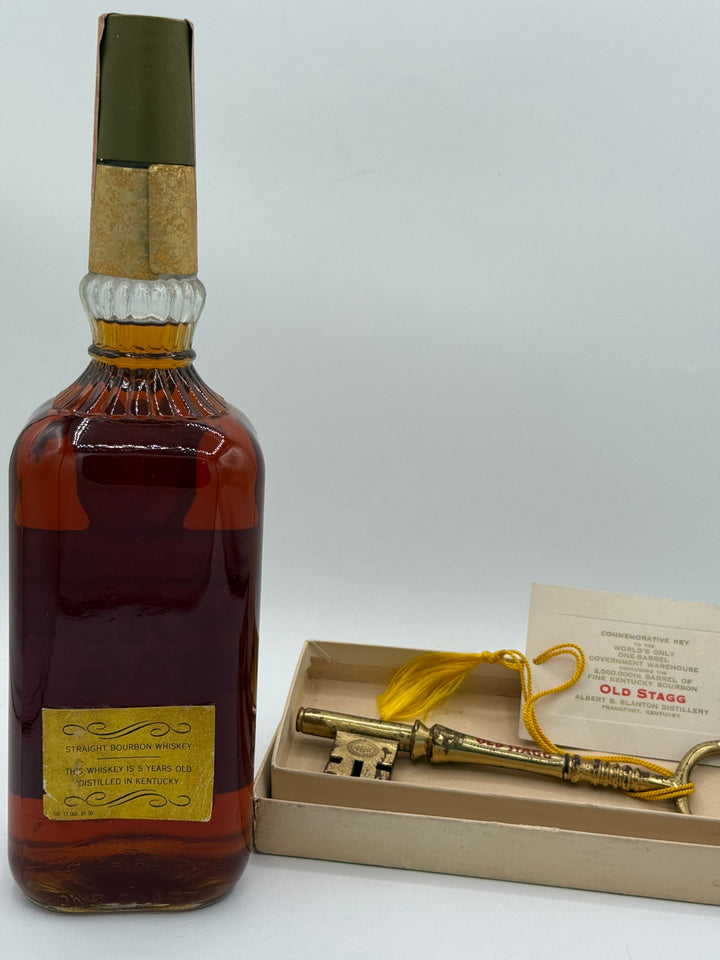 A vintage Old Stagg Straight Bourbon Whiskey bottle containing amber liquid with a yellow label is displayed alongside a gold commemorative key with a yellow tassel and an informational card detailing the World's Only One-Barrel Government Warehouse and the 2,000,000th barrel from the Albert B Blanton Distillery in Frankfort Kentucky