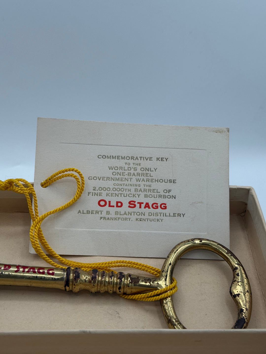 Photograph of an aged golden Old Stagg commemorative key inscribed June 23 1953, tied with a bright yellow cord to a white tag identifying it as for the 2,000,000th barrel of Old Stagg Kentucky Bourbon from the world's only one-barrel government warehouse at Albert B Blanton Distillery Frankfort Kentucky