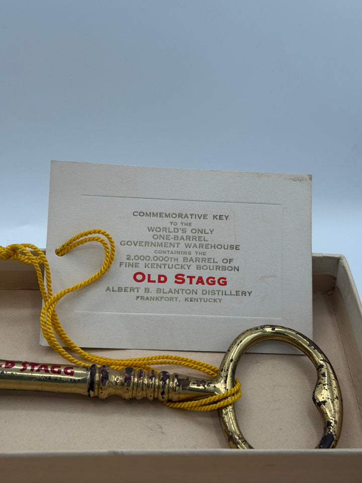 Photograph of an aged golden Old Stagg commemorative key inscribed June 23 1953, tied with a bright yellow cord to a white tag identifying it as for the 2,000,000th barrel of Old Stagg Kentucky Bourbon from the world's only one-barrel government warehouse at Albert B Blanton Distillery Frankfort Kentucky