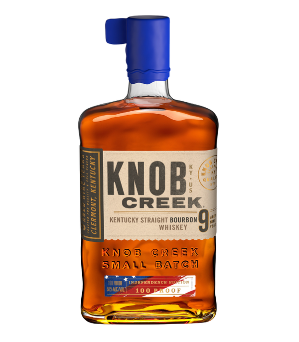 A clear glass bottle of Knob Creek Kentucky Straight Bourbon Whiskey, distilled in Clermont, Kentucky, featuring a dark amber liquid, a distinctive blue foil-wrapped cap with pull tab, a beige label displaying "Independence Edition" with a subtle American flag motif, and indicating "Aged 9 Years" and "100 Proof"