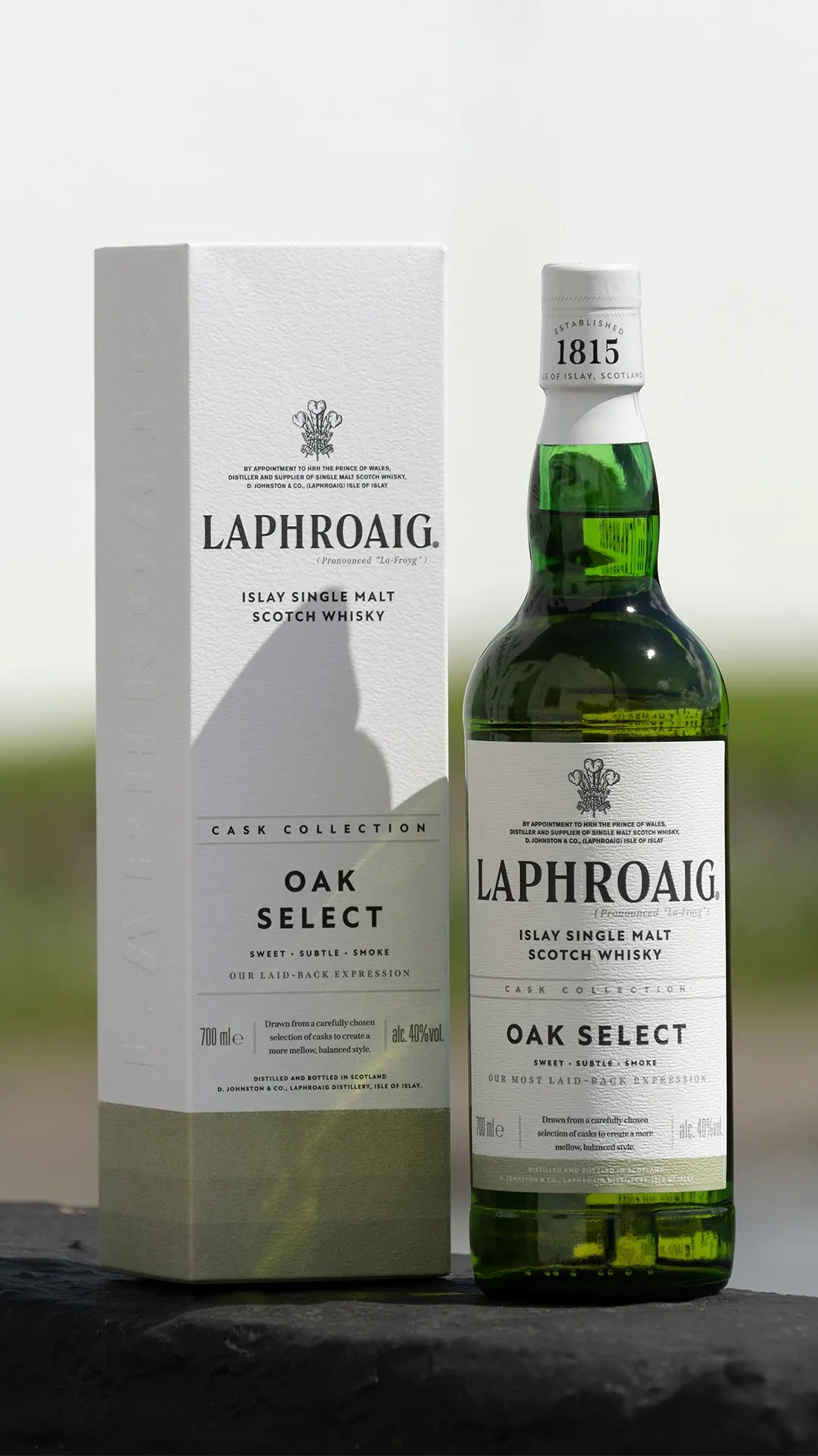 Photograph of a Laphroaig Islay Single Malt Scotch Whisky Oak Select green glass bottle and its matching white and olive-green box, featuring the "Established 1815" neck wrap and "By Appointment to HRH The Prince of Wales" crest, displayed outdoors on a dark stone surface
