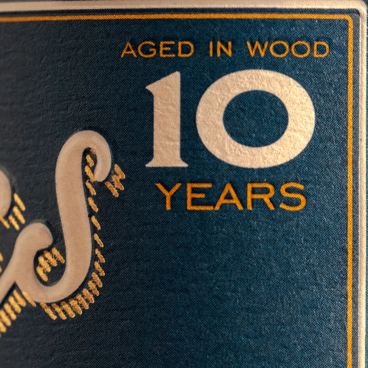 Photograph of a dark teal textured product label with prominent white number 10 and ornate letter S featuring gold dashed accents, gold text AGED IN WOOD and YEARS, and thin gold and white vertical border stripes