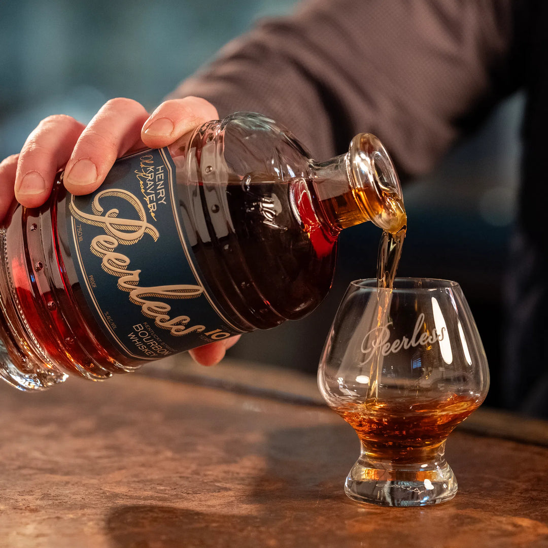 Photography of rich amber Peerless Kentucky Straight Bourbon Whiskey being poured from its distinctive dark blue and gold labeled bottle into a branded tasting glass on a warm bar counter