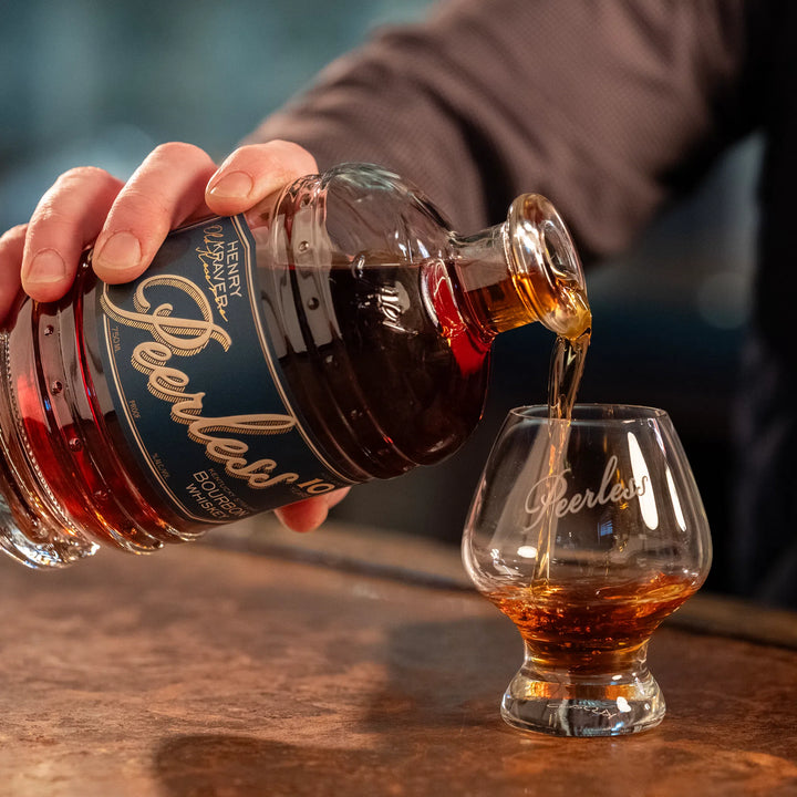 Photography of rich amber Peerless Kentucky Straight Bourbon Whiskey being poured from its distinctive dark blue and gold labeled bottle into a branded tasting glass on a warm bar counter