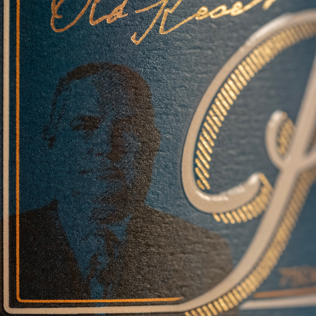 Close-up of a product label showing a dark silhouette of a man in a suit against a textured deep teal blue background with gleaming gold foil Old Reserve typography and an ornate initial G framed by a thin gold and white border