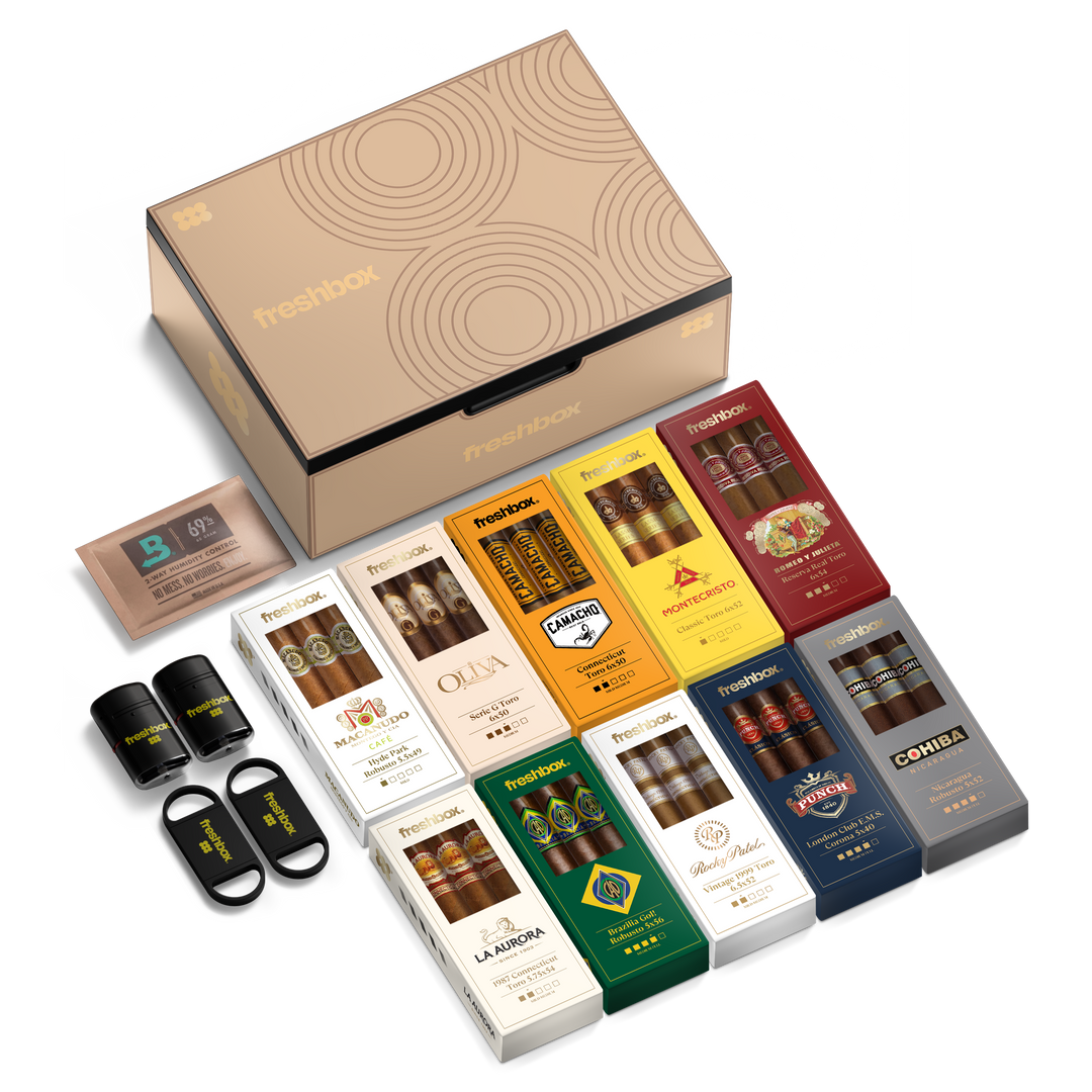 A studio product shot of a large beige Freshbox gift box featuring an array of twelve smaller colorful boxes of premium cigars from brands like Montecristo, Cohiba, and Macanudo, accompanied by black Freshbox cigar accessories and a brown Boveda humidity control pack