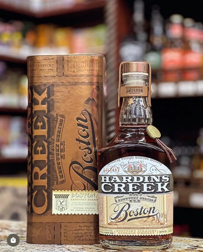 Photography of a Hardin's Creek Boston Kentucky Straight Bourbon Whiskey bottle and its ornate cylindrical presentation box on a store shelf, featuring amber liquid, a distinct label, a leather neck strap, and a gold medallion