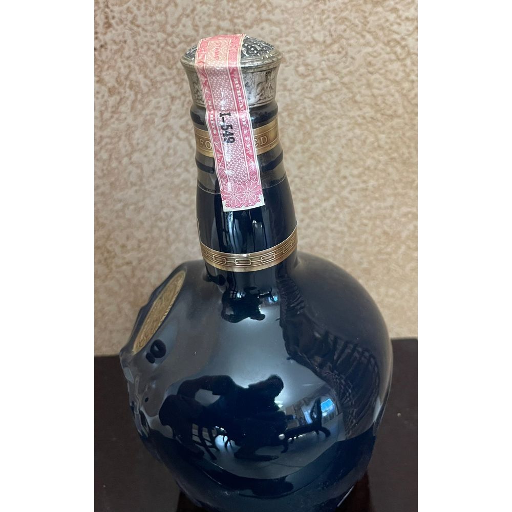 Product photography of a Royal Salute whisky bottle displayed indoors, featuring a glossy dark blue ceramic body with gold neck bands, an ornate silver stopper, and a distinctive pink tax stamp marked I-549